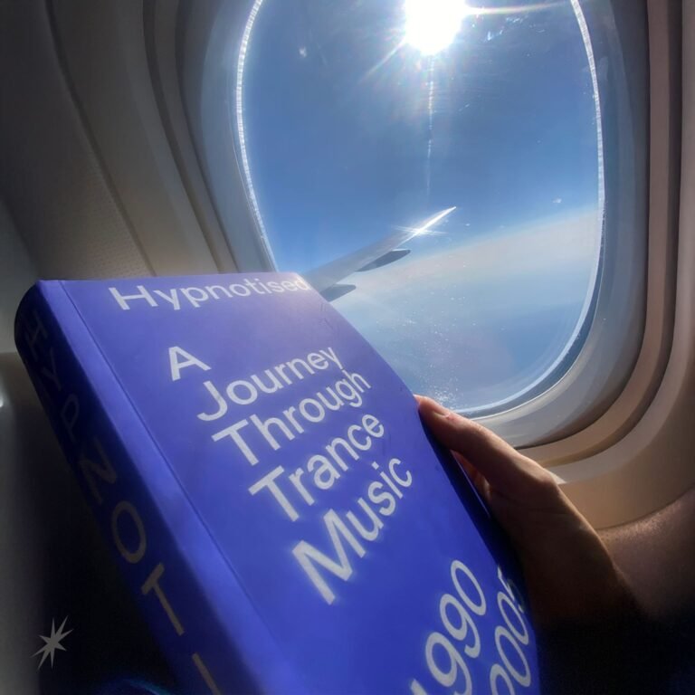 A person reads a bright blue book titled "Hypnotised: A Journey Through Trance Music" beside an airplane window showing a sunny sky and wingtip.