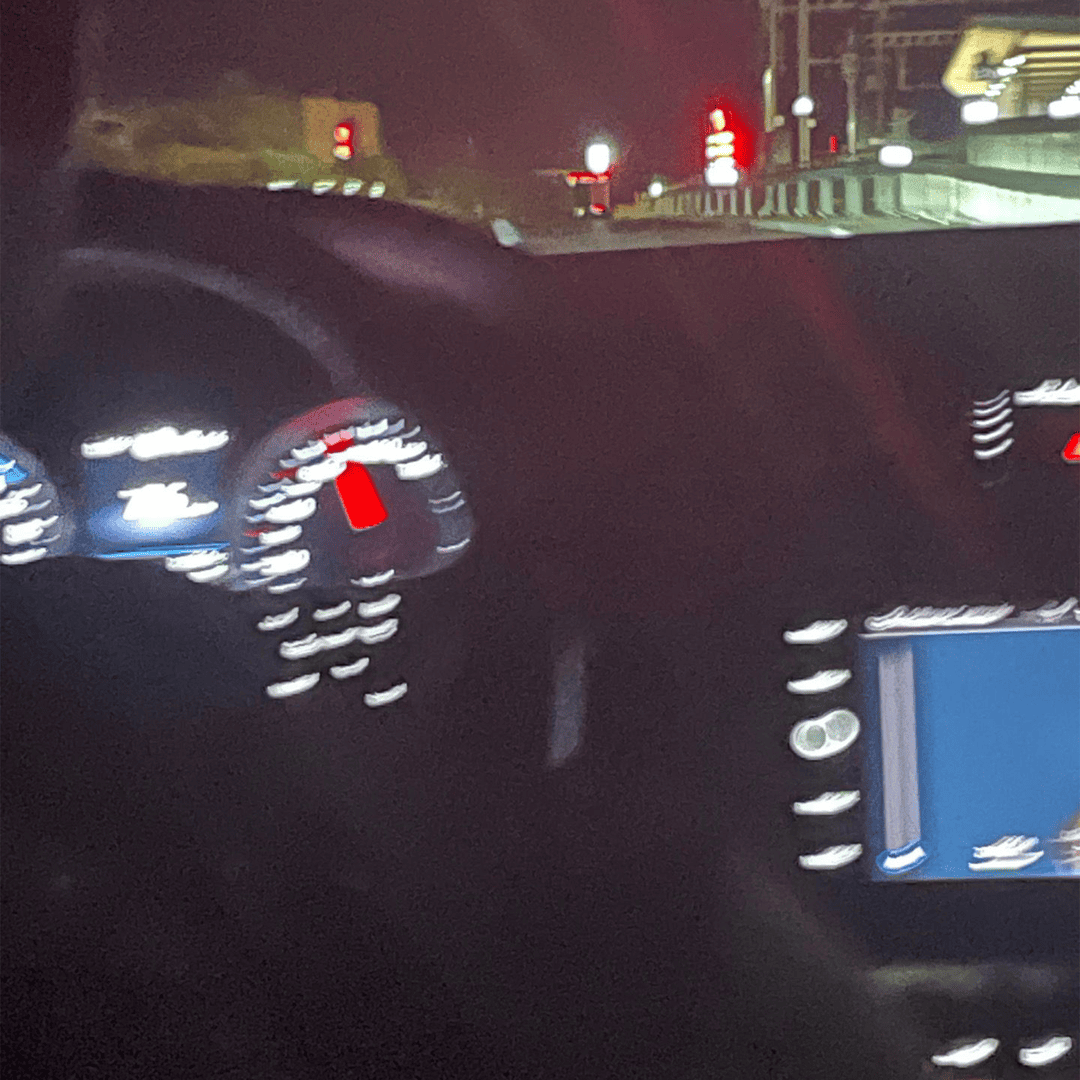 Blurry night view of a car dashboard with illuminated speedometer and digital display. Red traffic lights visible through the windshield.